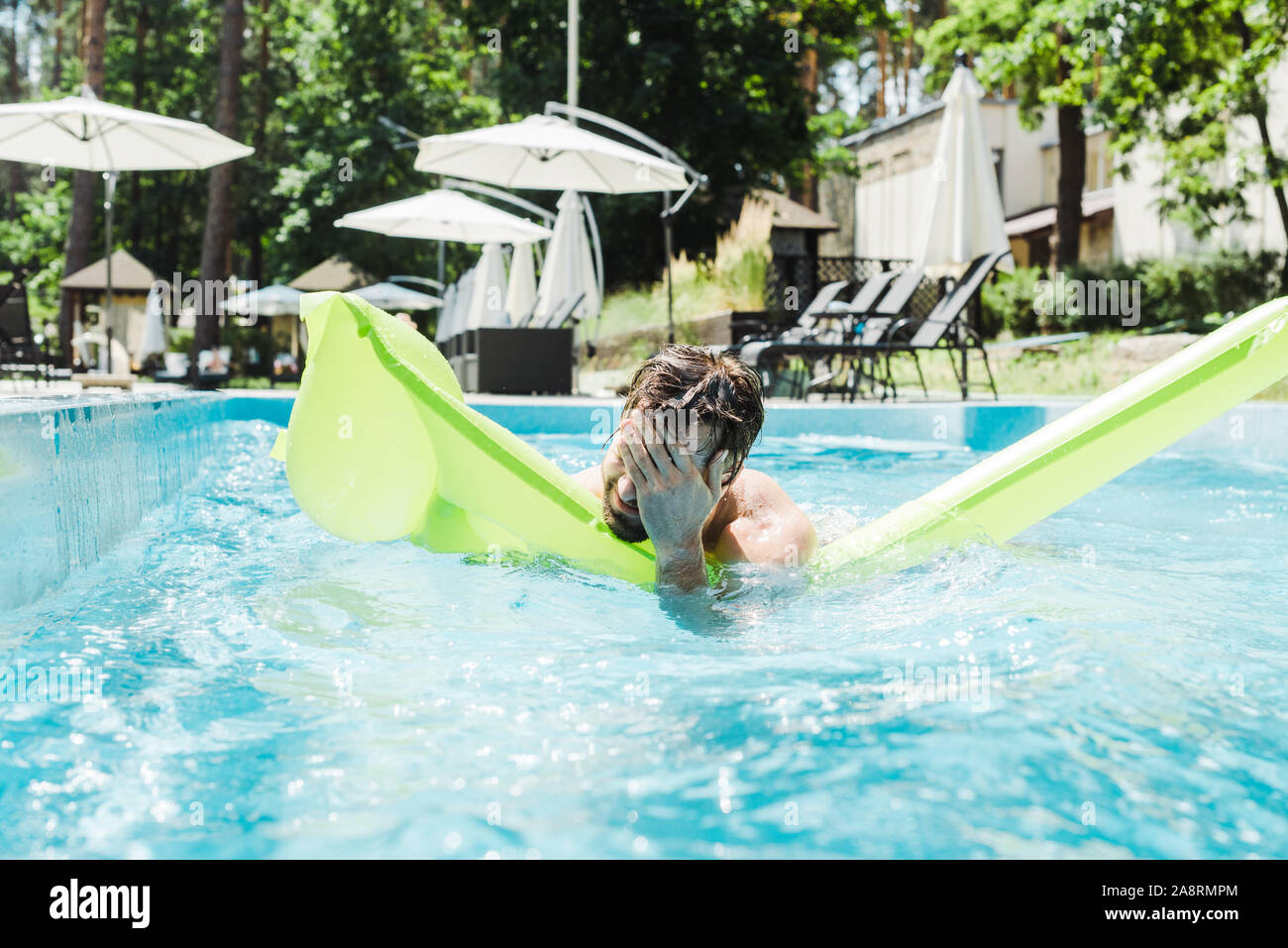 Uomo Barbuto di nuoto in piscina con verde anello gonfiabile e che copre il viso con le mani Foto Stock