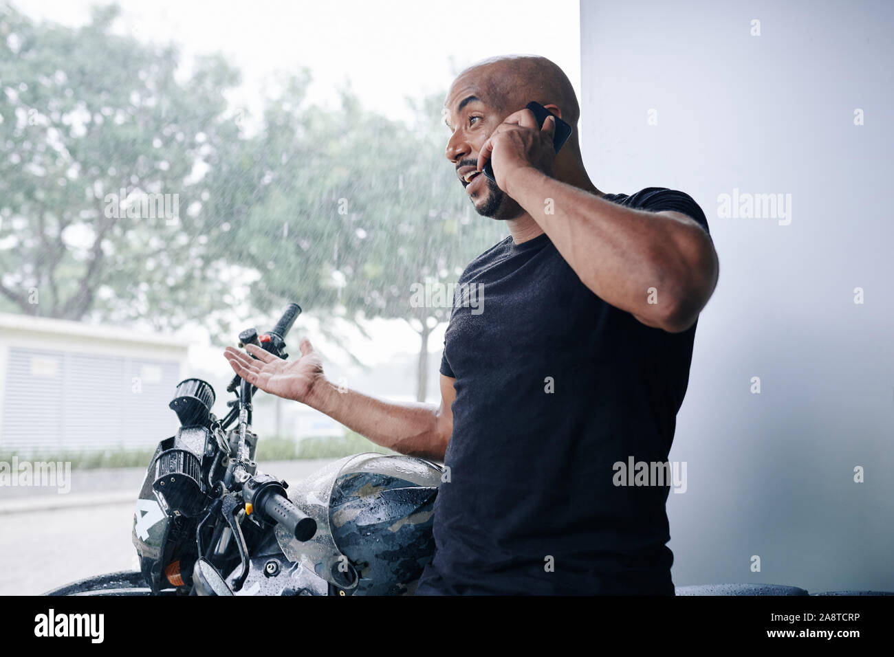 Uomo sorridente in piedi sotto il tetto con la sua bicicletta, chiamando il suo amico e lamenta di heavy rain Foto Stock