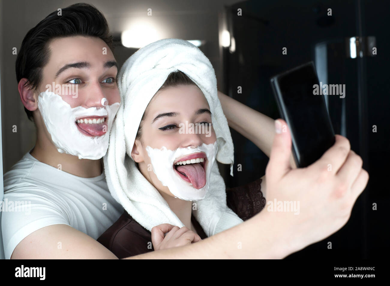 Funny giovane uomo bello e bella donna che colpisce una posa per selfie divertirsi sia con schiuma da barba sui loro volti in bagno allegro. Foto Stock