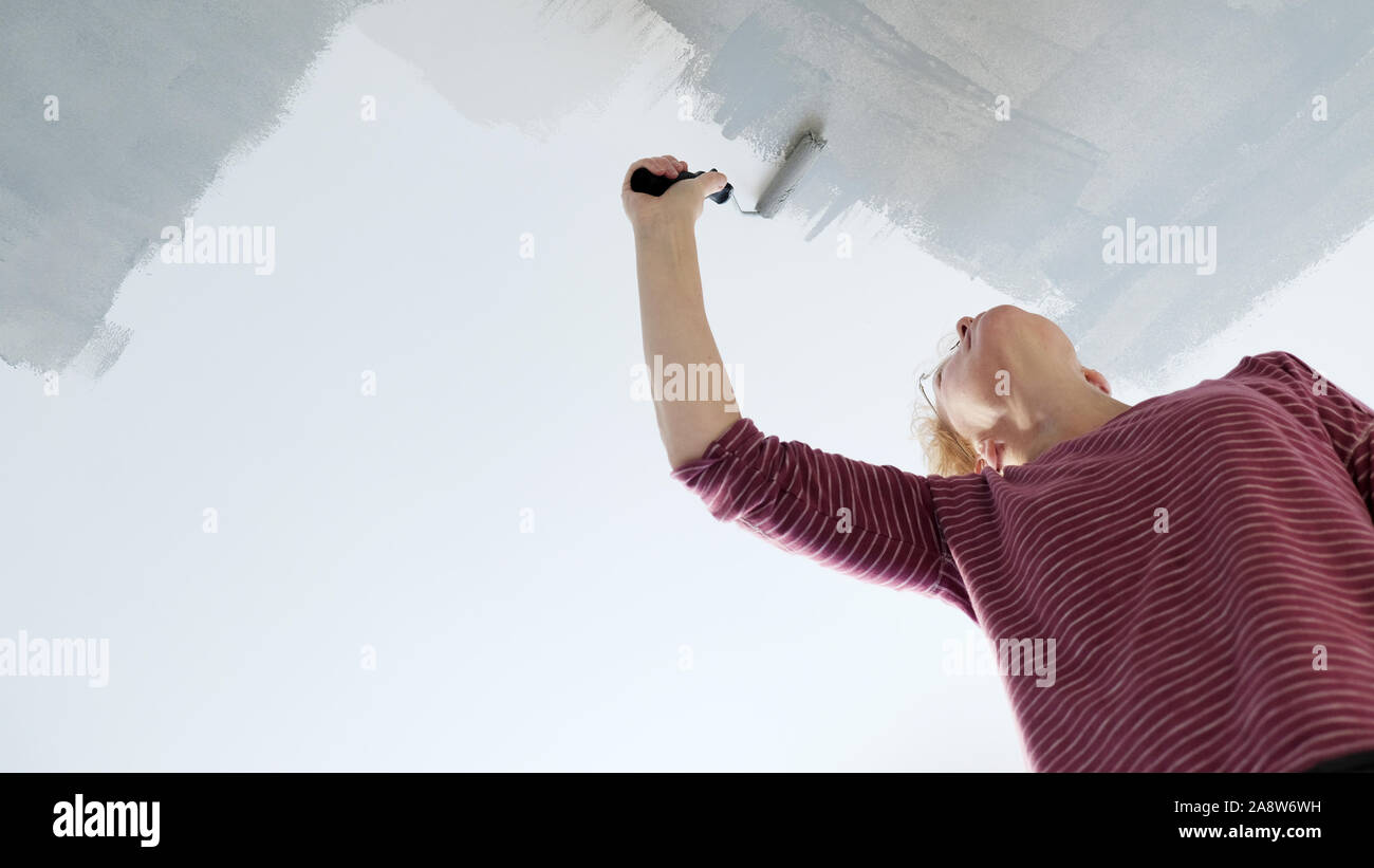 Una donna è la decorazione di casa sua, utilizzando un rullo di vernice per applicare una nuova vernice grigia su un soffitto bianco Foto Stock