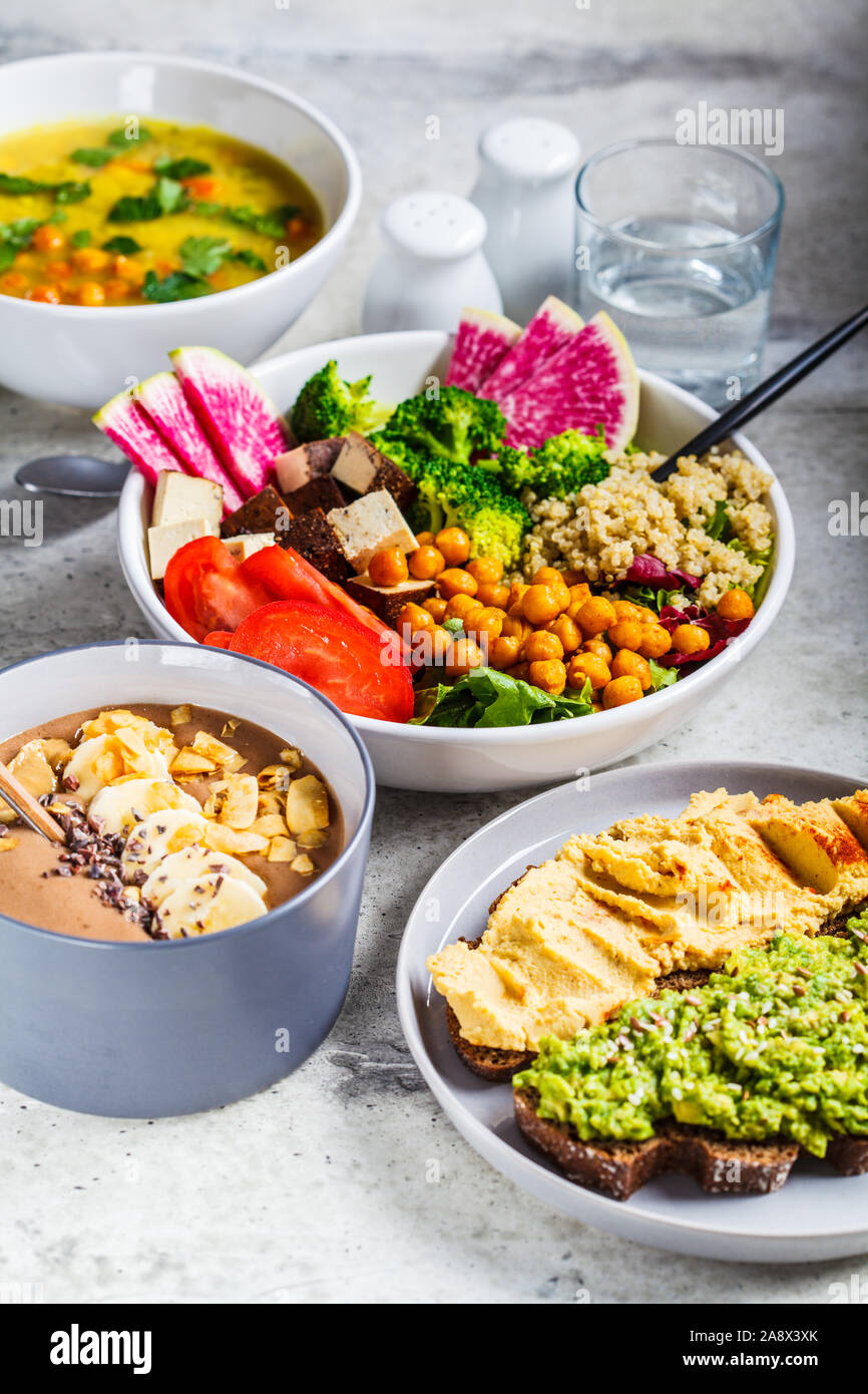 Vegan tavolo pranzo. Frullato al cioccolato ciotola, Buddha ciotola con tofu, ceci e la quinoa, zuppa di lenticchie e toast su sfondo grigio. Foto Stock