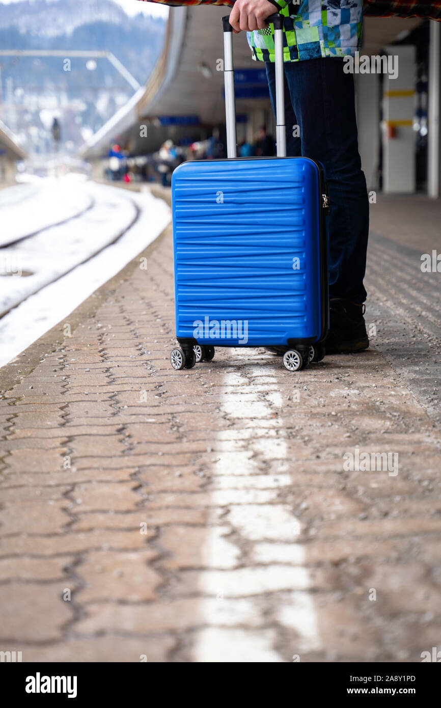 Turistico con valigia blu alla stazione in attesa del treno contro sullo sfondo di montagne sulla giornata invernale Foto Stock
