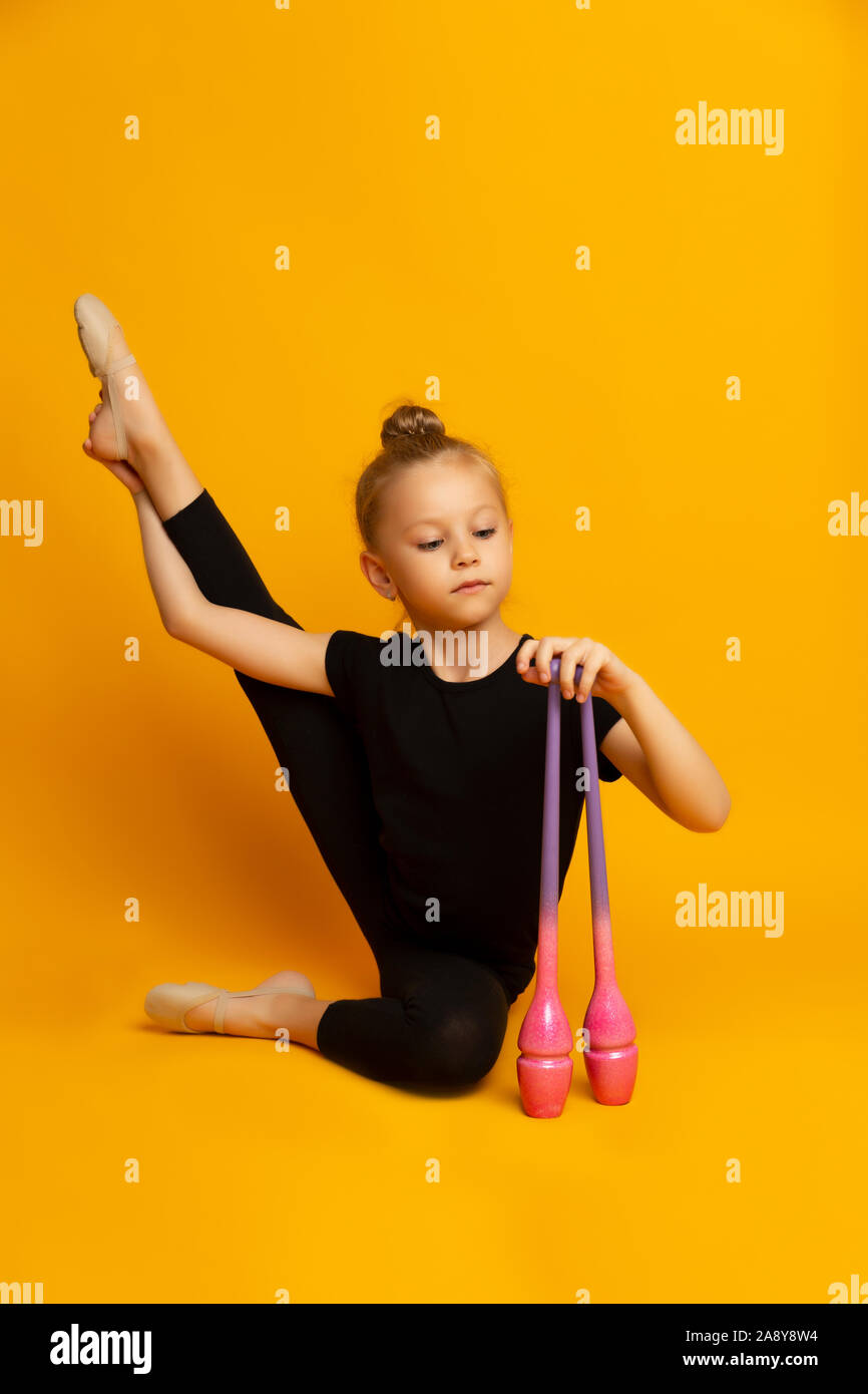 Bambina in un costume nero si allunga la gamba e guarda verso il basso, e trattiene i club sportivi in mano Foto Stock