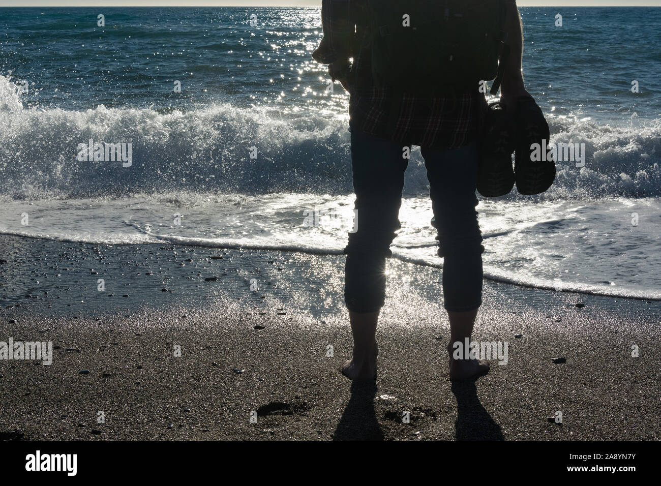 Un uomo a piedi nudi che porta un piccolo zaino in piedi su una spiaggia tropicale con le sue scarpe nelle sue mani, retroilluminato e guardando nel sole Foto Stock