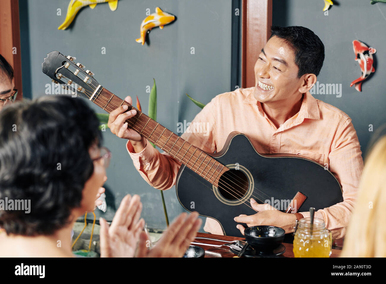 Felice di mezza età uomo asiatico a suonare la chitarra e cantare una canzone per i suoi parenti a cena in famiglia Foto Stock