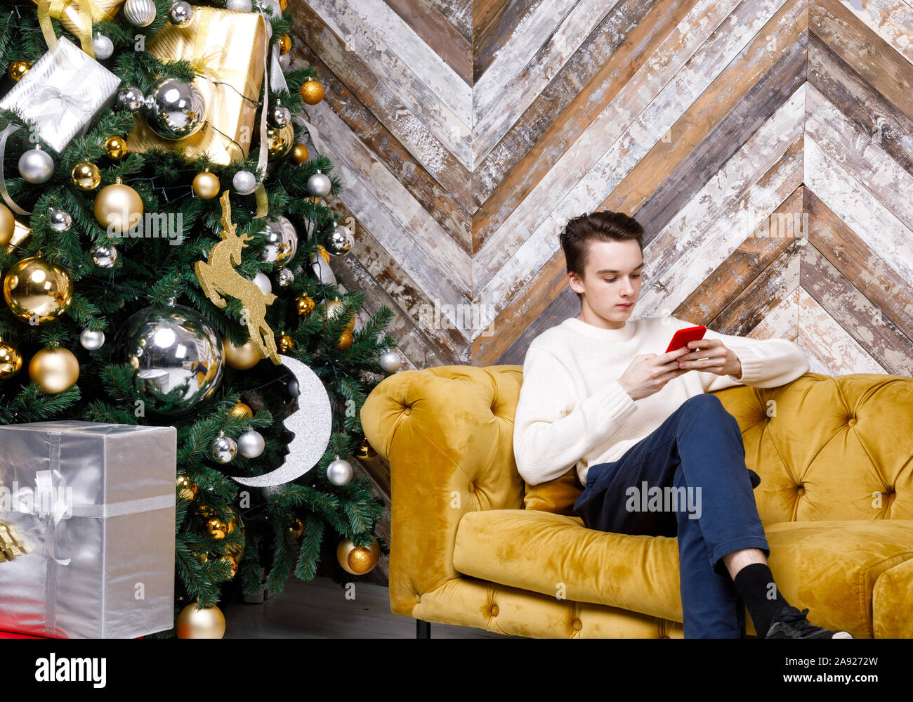Adolescente ragazzo seduto da solo su un divano accanto a Albero di Natale si è concentrato solo sul suo smartphone messaging o il controllo di social media - vacanza Smombie concep Foto Stock