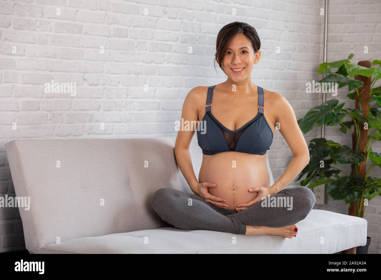 Giovani asiatici donna incinta aspettano di nuovo bambino si siede sul divano di casa. Ella è di indossare abbigliamento sportivo e guardando la fotocamera. Foto Stock