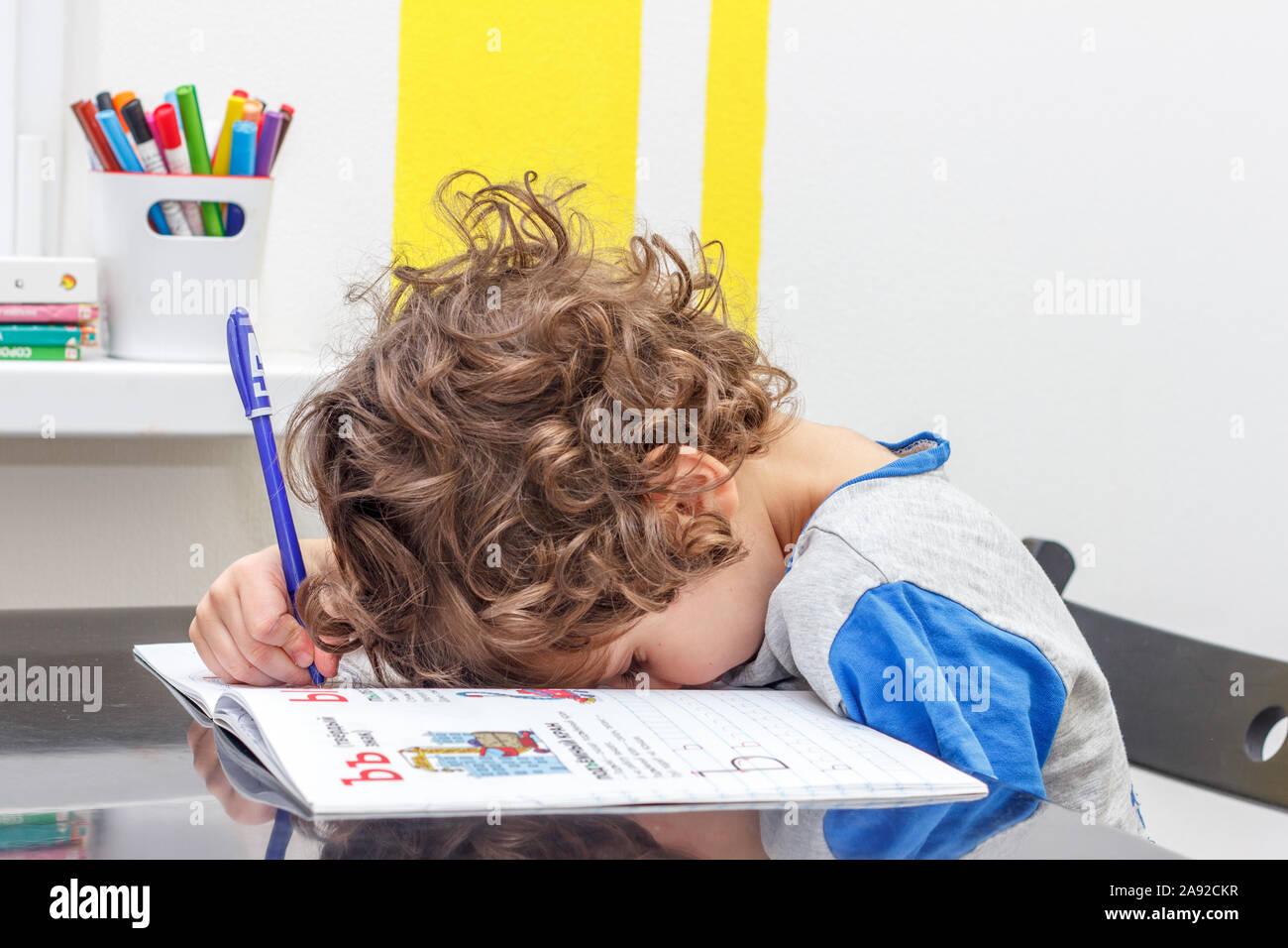 Bambino stanco seduto al tavolo, scrivendo nel suo notebook mentre fa i compiti. Concetto di un gran numero di incarichi a scuola Foto Stock