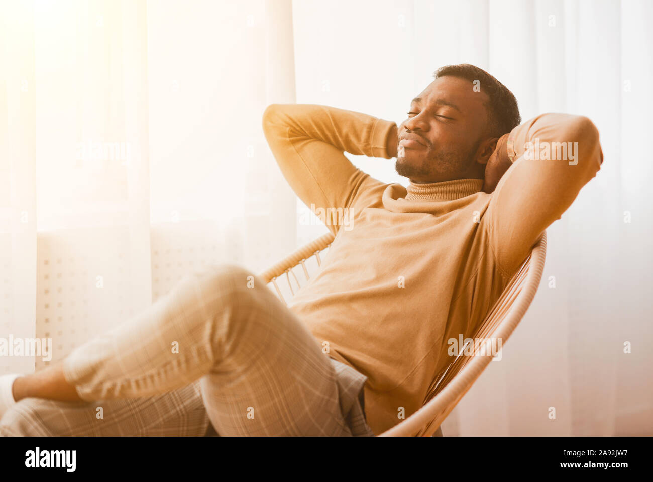 Giorno pigro a casa. Afro uomo rilassante tenendo le mani dietro la testa seduto sulla sedia moderna accanto alla finestra Indoor. Foto Stock