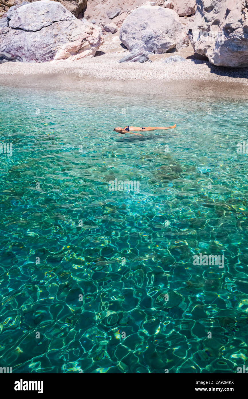 Donna turistica che galleggia sulle sue spalle sulle acque cristalline e turchesi della Baia di Galazia Nera; Isola di Polyaigos, Cicladi, Grecia Foto Stock