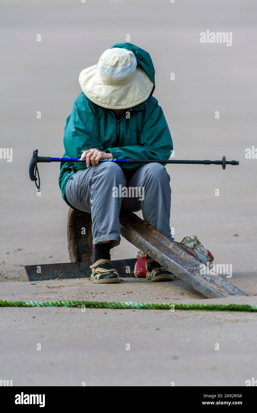 Femmina con la faccia nascosta dal cappello beige, seduto sul dispositivo di ancoraggio sulla spiaggia, riposo durante una passeggiata, indossa una giacca verde, blu jeans e portante Walking palo. Foto Stock
