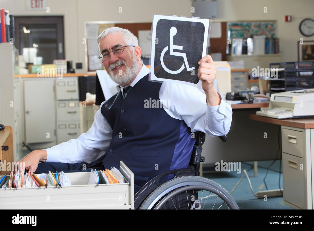 Uomo con distrofia muscolare in una sedia a rotelle che lavora in un ufficio e tenendo in mano un cartello per portatori di handicap Foto Stock