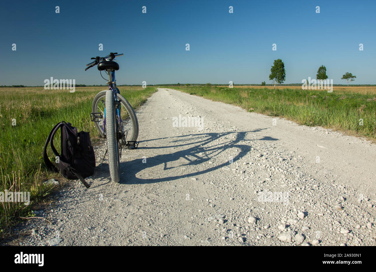 Bicicletta permanente e uno zaino su una strada di ghiaia all'orizzonte, prato verde, alberi e cielo blu senza nuvole Foto Stock