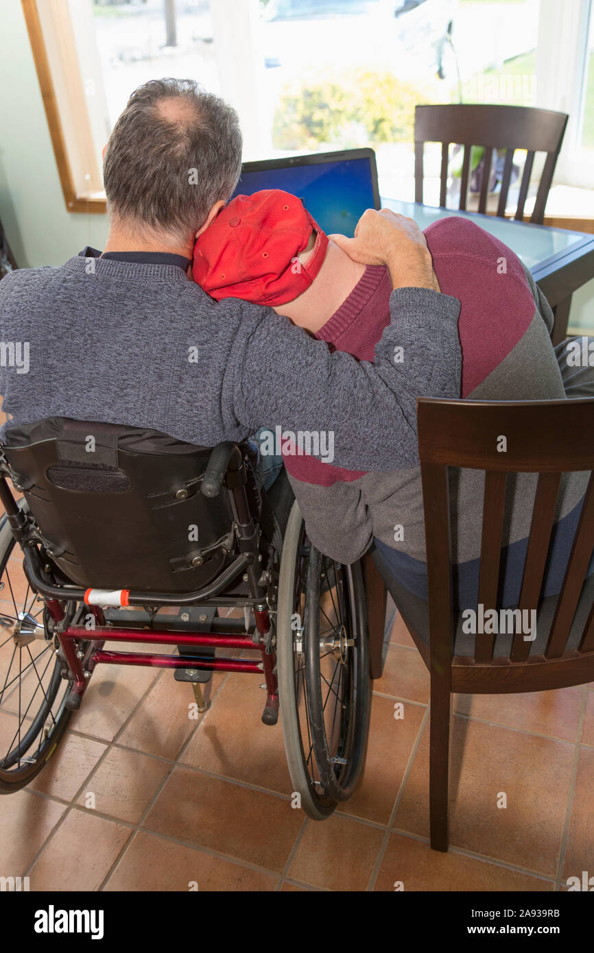 Giovane uomo con sindrome di Down abbracciando suo padre in sedia a rotelle Con lesione del midollo spinale a casa Foto Stock