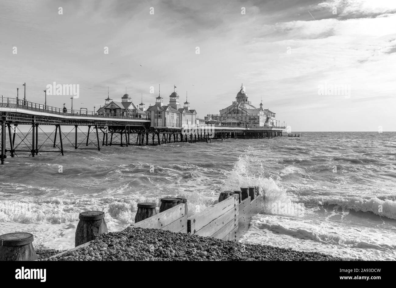 La mattina presto a una spiaggia di ciottoli a Eastbourne. Un'onda rompe contro una scogliera di legno in primo piano e un molo è al di là. Foto Stock