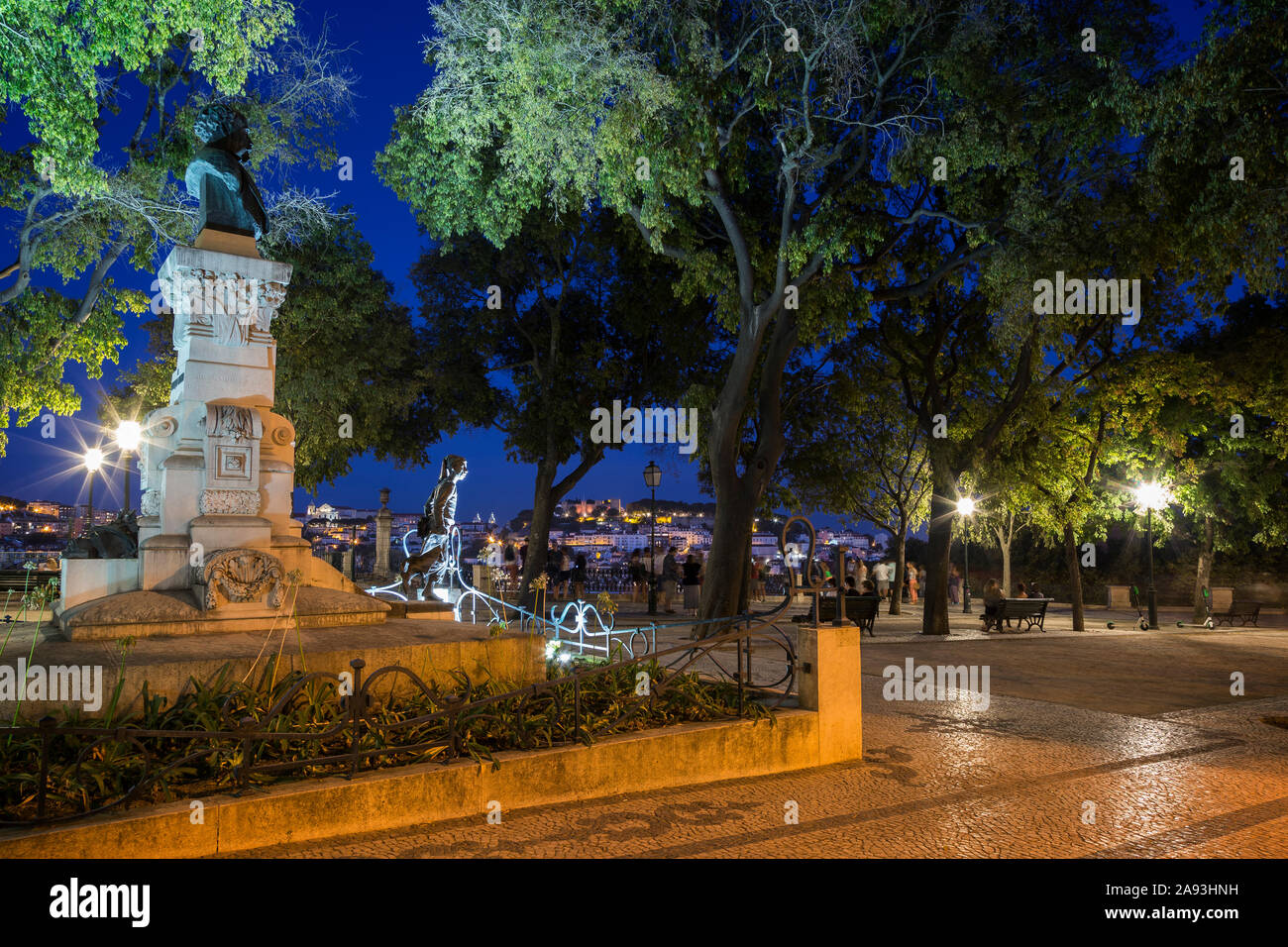 Statua illuminata e persone in un parco al Miradouro de Sao Pedro de Alcantara viewpoint a Lisbona, Portogallo, la sera. Foto Stock