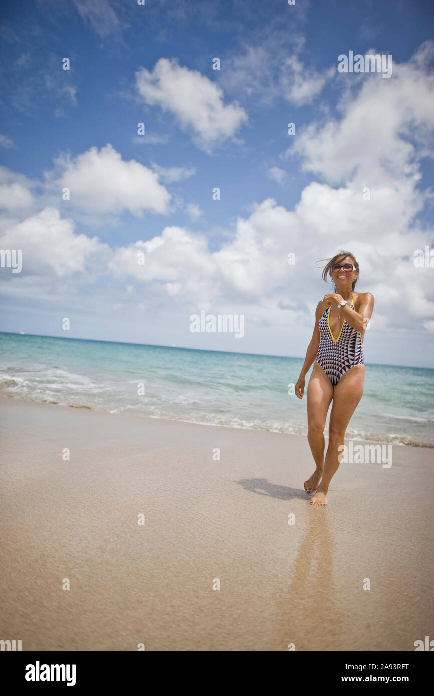 Sorridente metà donna adulta camminando lungo una spiaggia nel suo costume da bagno e occhiali da sole. Foto Stock