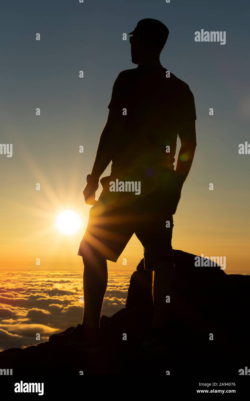 Uomo silhoueted che si erge sulla roccia sopra le nuvole al tramonto, Haleakala; Maui, Hawaii, Stati Uniti d'America Foto Stock