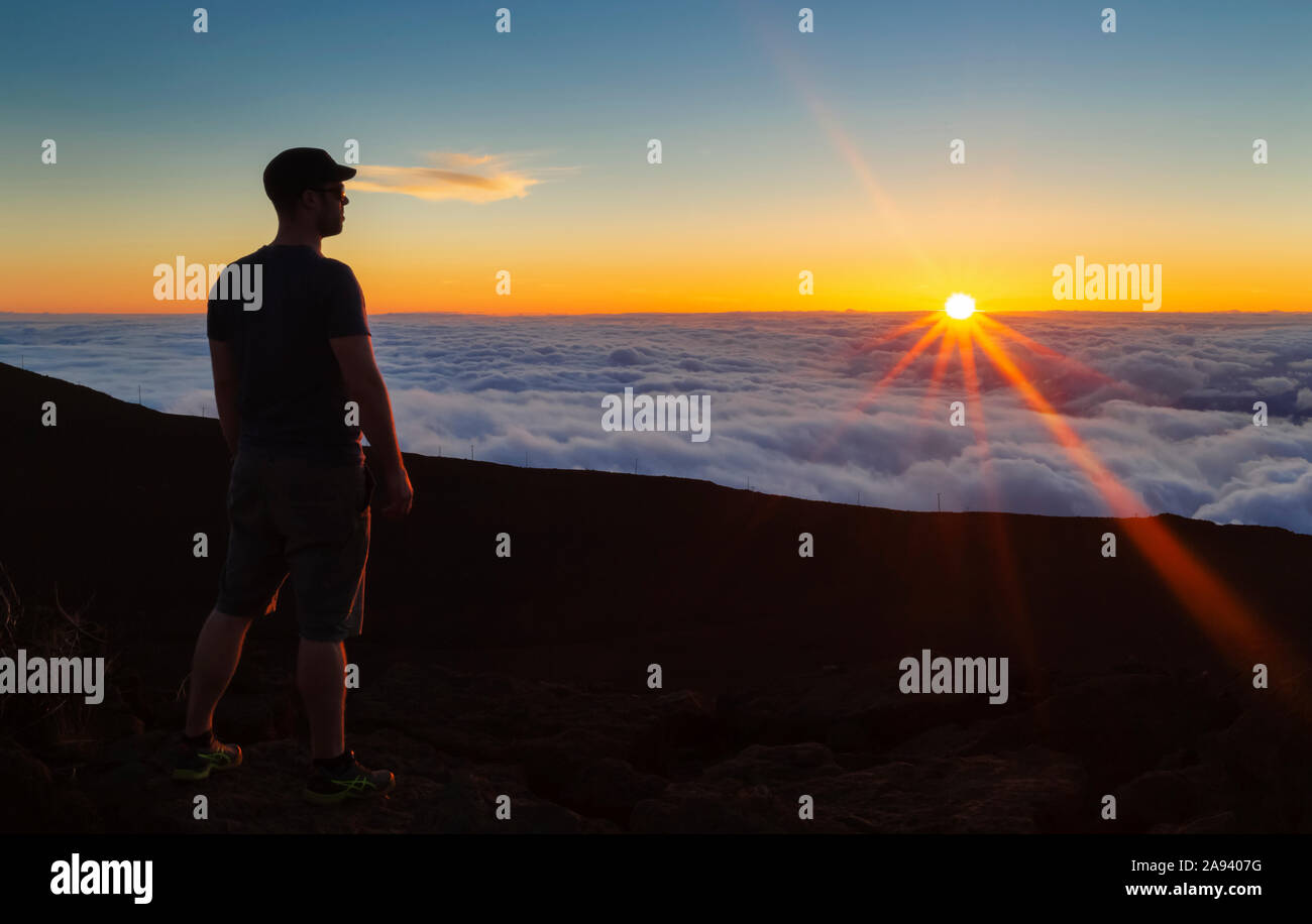 Uomo silhoueted che si erge sulla roccia sopra le nuvole al tramonto, Haleakala; Maui, Hawaii, Stati Uniti d'America Foto Stock