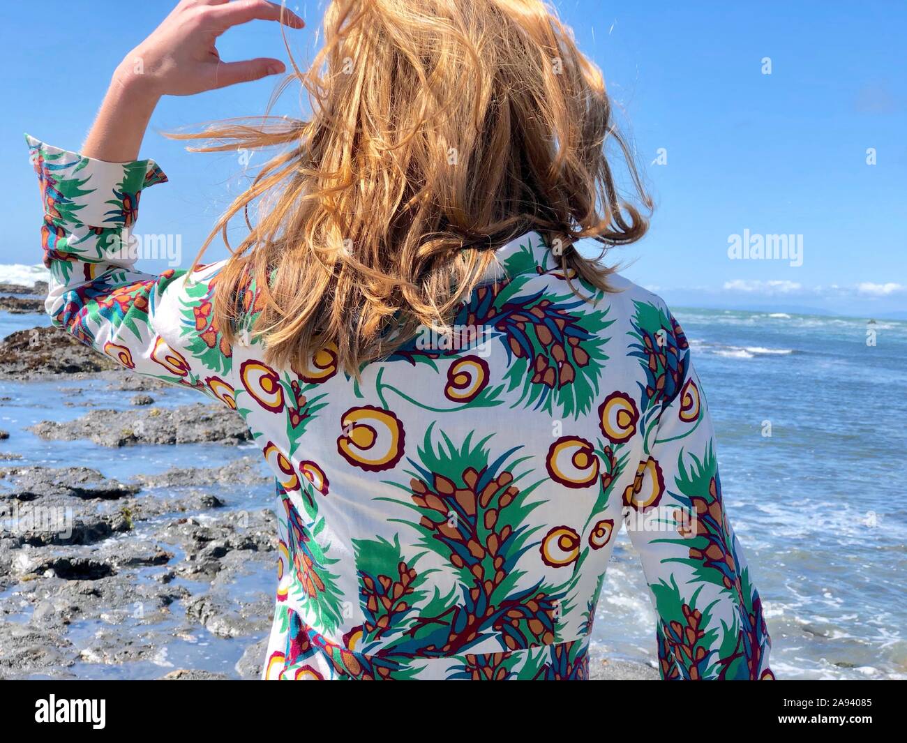 Giovani rossastro-donna bionda visto da dietro, indossando abiti floreali, il vento nei capelli, a guardare il mare dalla riva Foto Stock