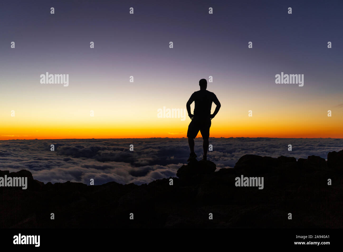 Uomo silhoueted che si erge sulla roccia sopra le nuvole al tramonto, Haleakala; Maui, Hawaii, Stati Uniti d'America Foto Stock