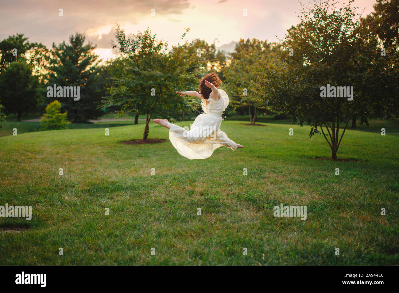 Un ballerino salta in un lungo abito bianco in un parco alberato al tramonto Foto Stock