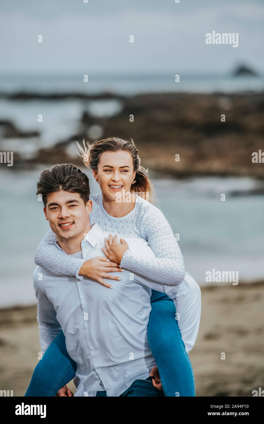 Ritratto di una giovane coppia in spiaggia, la giovane donna che viene portata sulla schiena del giovane; Wellington, Isola del Nord, Nuova Zelanda Foto Stock