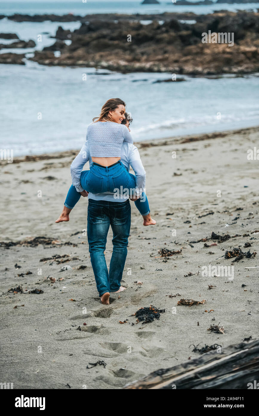 Una giovane coppia in spiaggia, la giovane donna che viene portata sulla schiena del giovane uomo; Wellington, Isola del Nord, Nuova Zelanda Foto Stock