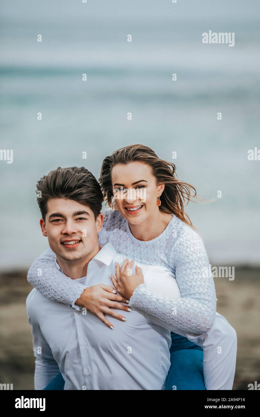 Ritratto di una giovane coppia in spiaggia, la giovane donna che viene portata sulla schiena del giovane; Wellington, Isola del Nord, Nuova Zelanda Foto Stock