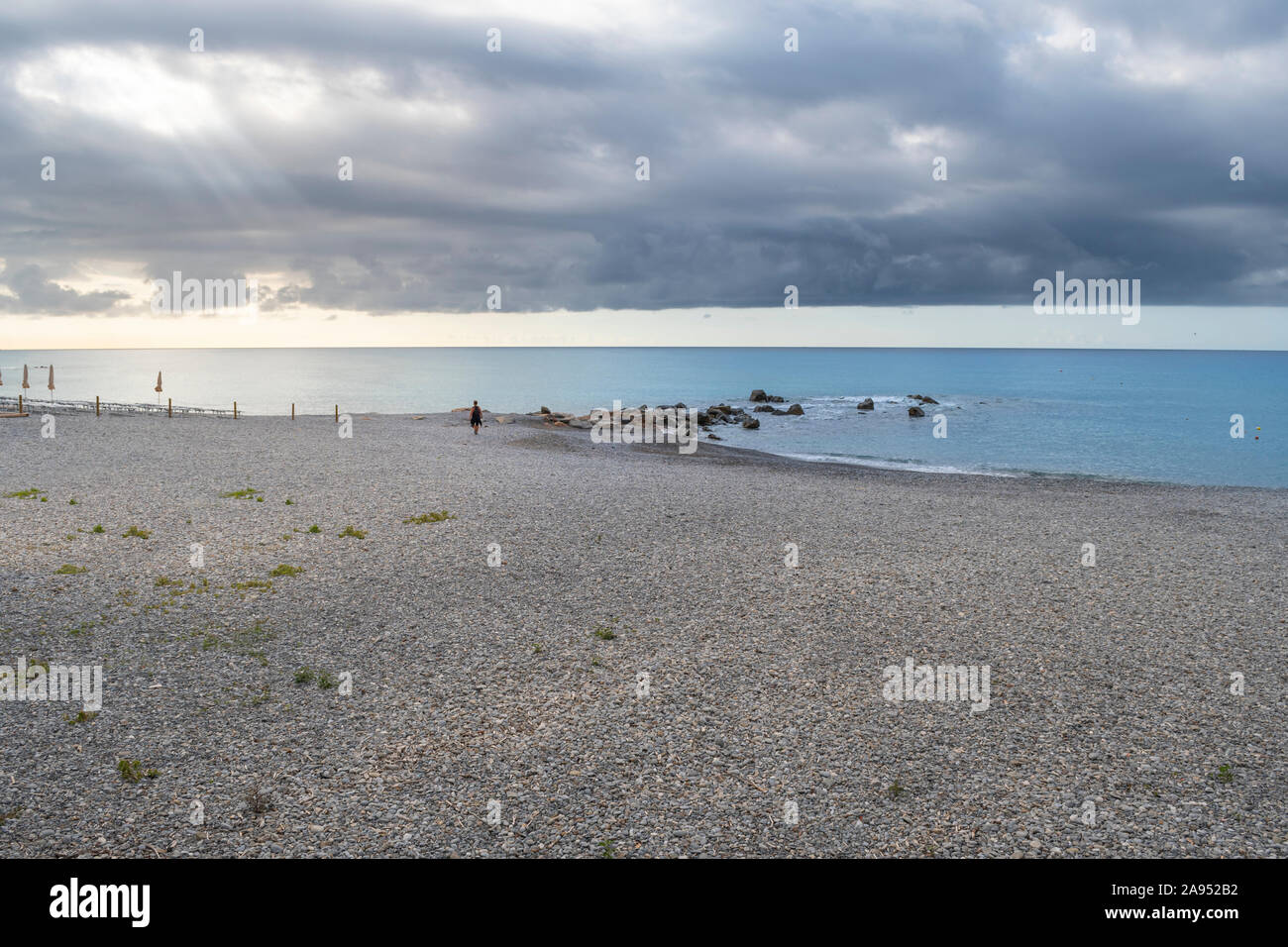 Un identificabili lone uomo cammina lungo il litorale di Ventimiglia, Italia su un mattino nuvoloso come i raggi del sole il picco attraverso le nuvole Foto Stock