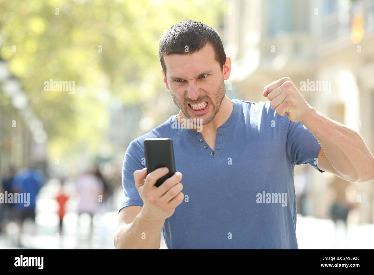 Vista frontale ritratto di un uomo arrabbiato controllo si è schiantato smart phone in piedi in strada Foto Stock
