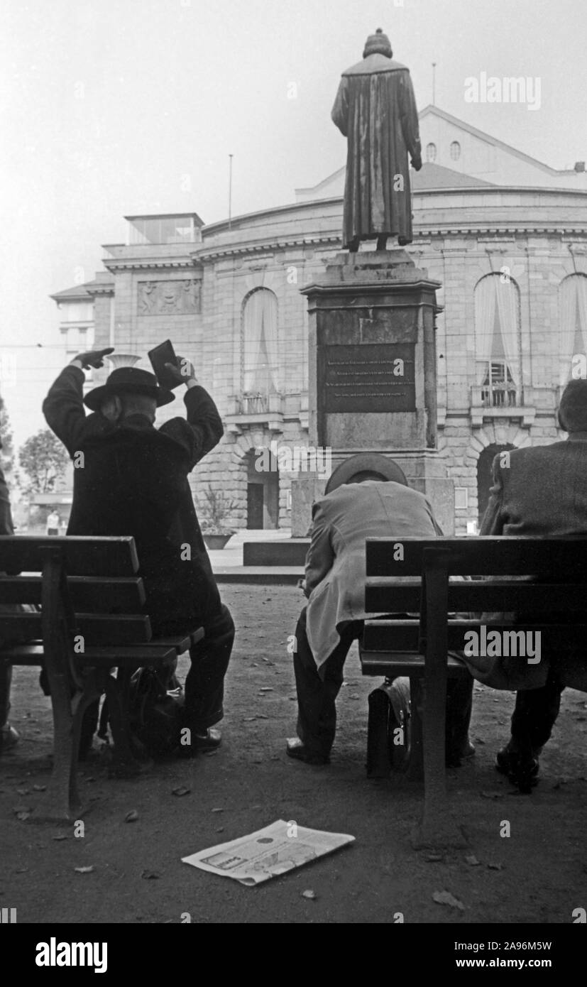 Mainzer sitzen auf Bänken auf dem Gutenbergplatz und blicken auf das Gutenbergdenkmal und das Stadttheater in Mainz, Deutschland 1961. Abitanti di Magonza seduti sui banchi sulla Gutenbergplatz square, rivolto verso la parte posteriore del monumento di Gutenberg e la città del teatro, Germania 1961. Foto Stock