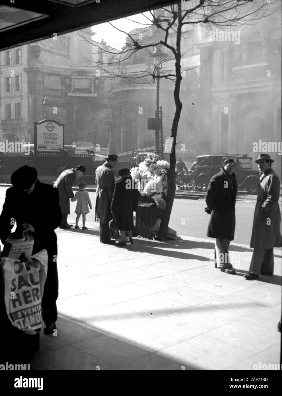 Un venditore di giornali e fiori che lavora la strada alla Holborn Station di Londra nel 1952. Una vita quotidiana 1950s Street scene con auto veterane in background. Foto Stock