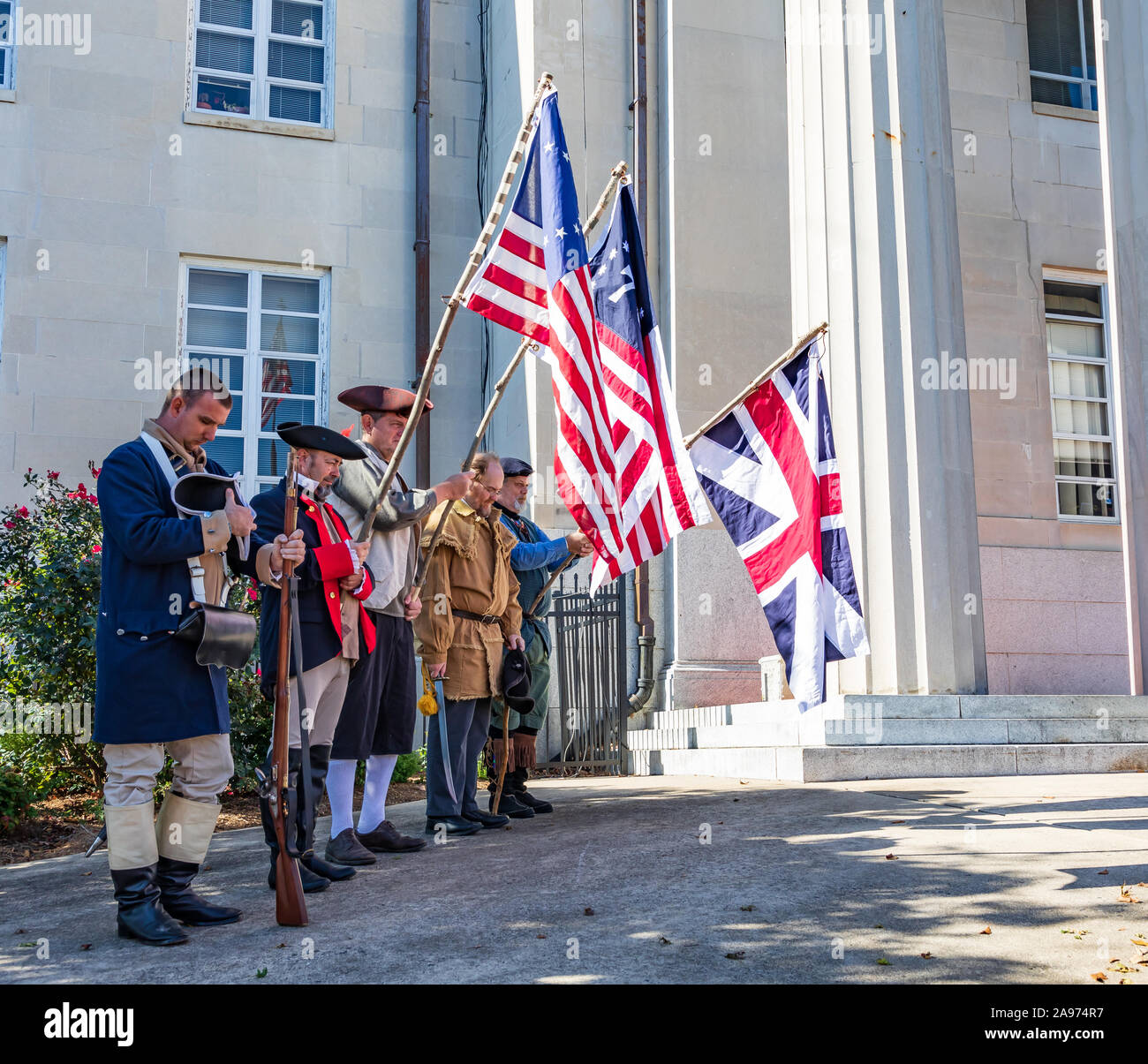 LINCOLNTON, NC, Stati Uniti d'America-11 NOV 2019: una linea di uomini vestiti in periodo negli Stati Uniti costume militare, tenendo bandiere storiche, con testa inchinata nella preghiera. Foto Stock