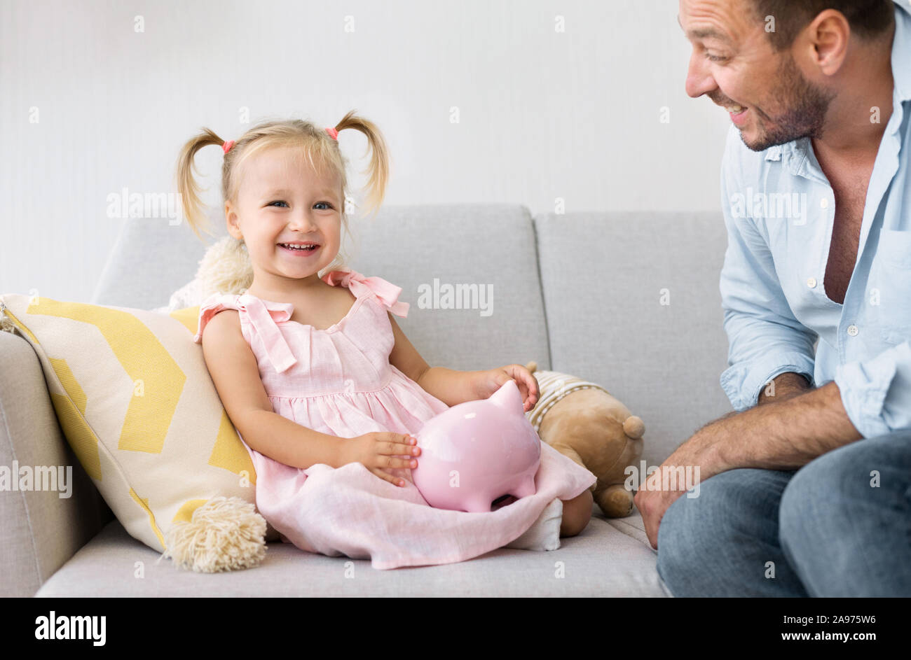 Bella ragazza seduta con Piggybank, giocando con il papà sul lettino, guardando la fotocamera Foto Stock