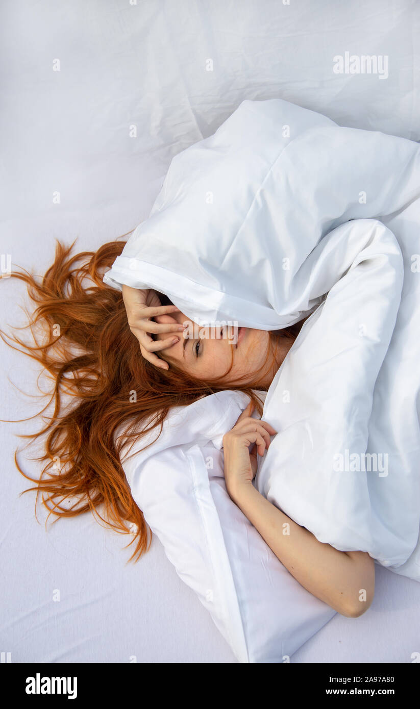 Attraente, giovani, i capelli rossi donna, capelli wild sui fogli, faccia metà sotto il cuscino giacente in fresco morbido fogli bianchi in camera da letto, spazio di copia Foto Stock