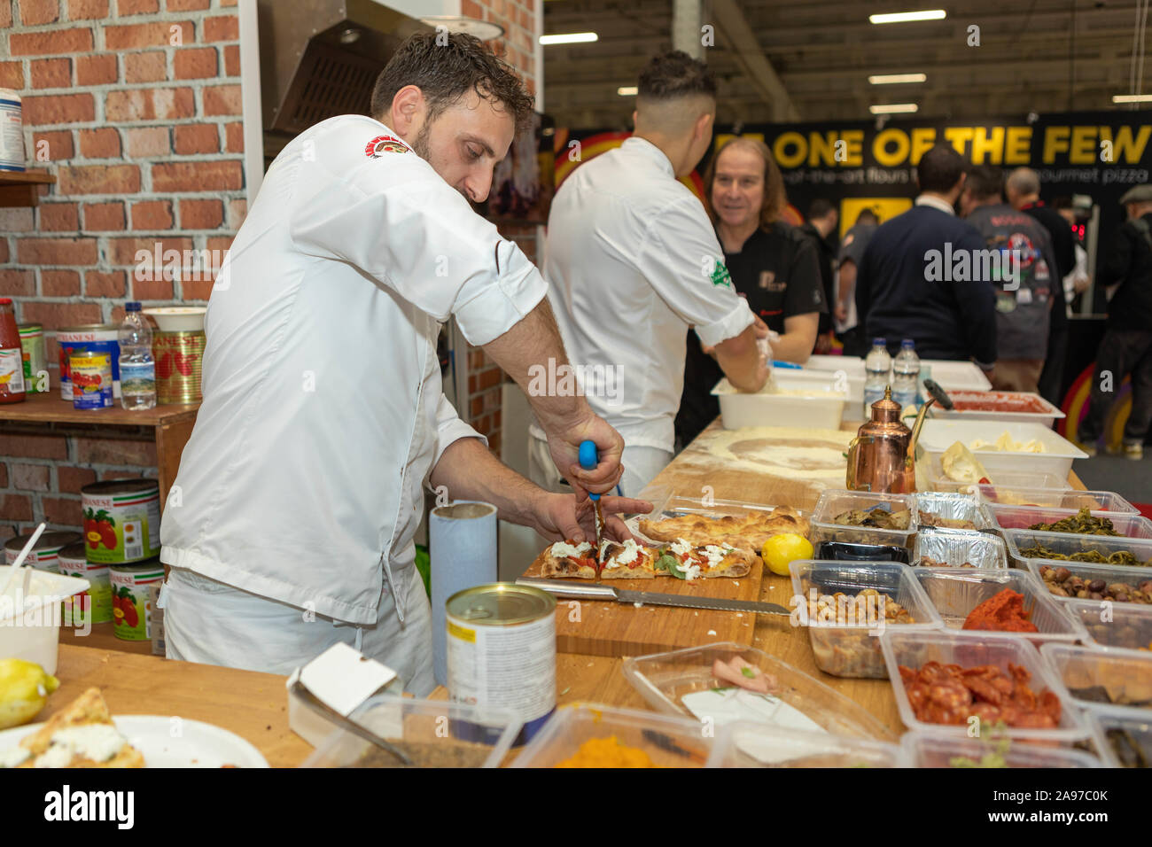 Olympia, Londra, Regno Unito. 13 novembre, 2019. Uno chef affetta una pizza appena fatta usando un tagliapasta in un evento culinario pubblico. La tavola è piena di ingredienti freschi, tra cui funghi, pomodori ciliegini, limoni e prodotti in scatola. Altri chef stanno preparando il cibo in sottofondo. I principali grossisti di prodotti alimentari del Regno Unito e dell'Europa partecipano alla fiera Pizza and Pasta food. Penelope Barritt/Alamy Live News Foto Stock