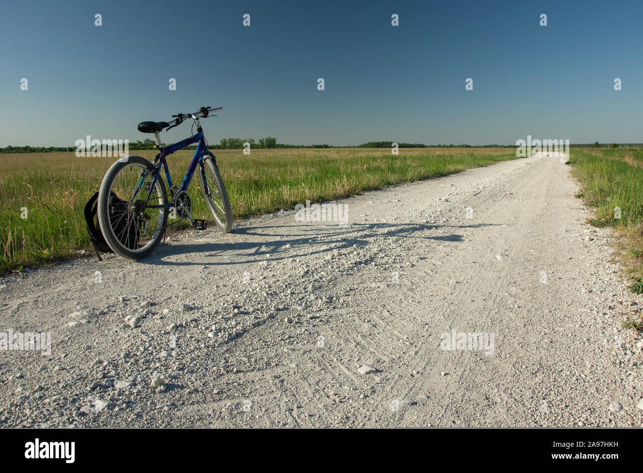 Bicicletta permanente e uno zaino su una strada di ghiaia all'orizzonte, prato verde e cielo blu senza nuvole Foto Stock