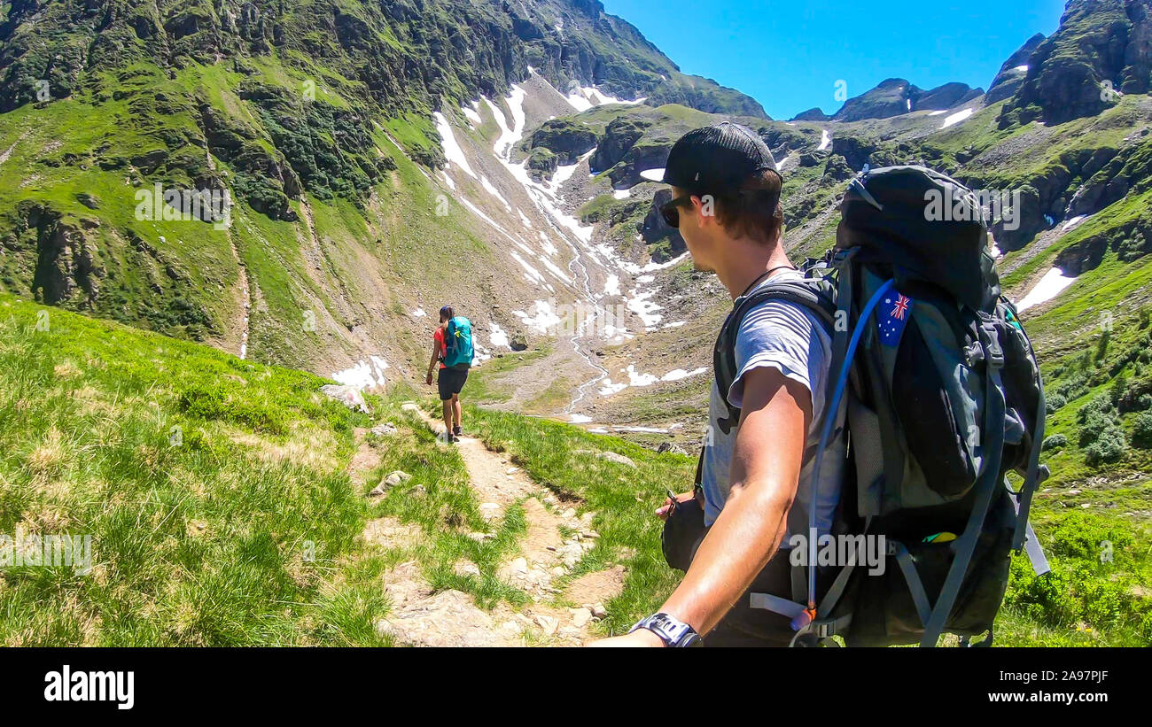 Un giovane con un grande zaini escursioni verso il basso su un ripido sentiero tra alti picchi di montagna. Alcune delle piste sono coperti di neve. Nella parte posteriore è anot Foto Stock