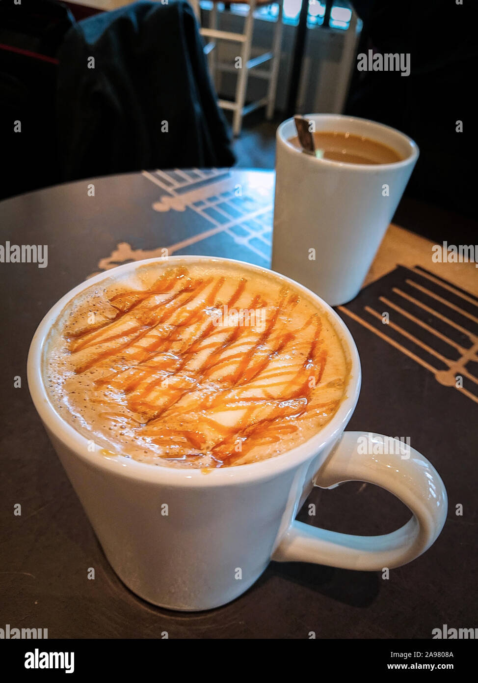 Bianco tazza ceramica riempito di caffè e una bella decorata crema sulla sommità in piedi su una tavola di marrone con una seconda tazza in background Foto Stock Bianco tazza ceramica riempito di caffè e una bella decorata crema sulla sommità in piedi su una tavola di marrone con una seconda tazza in background Foto Stock