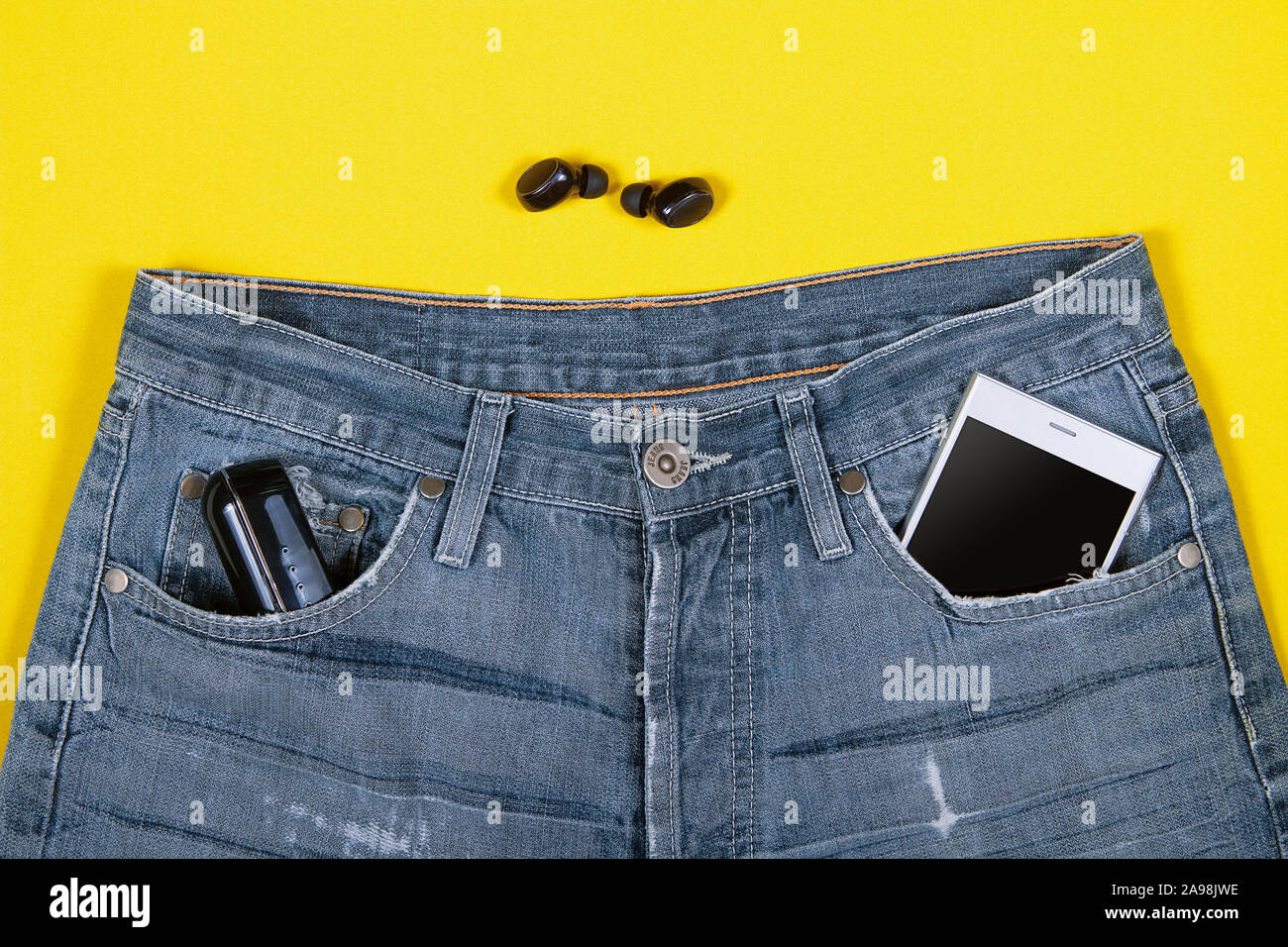 Vista dall'alto di cuffie wireless, jeans, caricabatteria e lo smartphone nella sua tasca su sfondo giallo Foto Stock