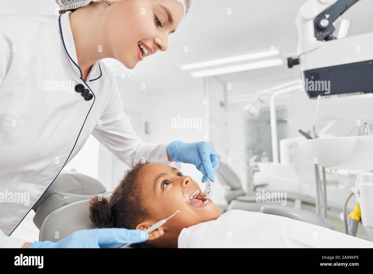 Dentista femmina in camice bianco e guanti di gomma il trattamento di denti della piccola ragazza africana. Paziente seduto in poltrona odontoiatrica con bocca aperta. Medico azienda strumenti dentali e guardando i denti. Foto Stock
