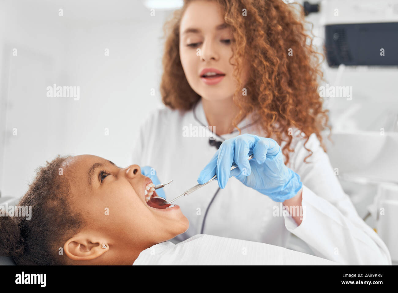 Bellissima femmina dentista di ispezionare i denti del piccolo paziente africano. Dottore in camice bianco e guanti di gomma holding strumenti dentali e sta esaminando la bocca della ragazza. Concetto di cure dentarie. Foto Stock