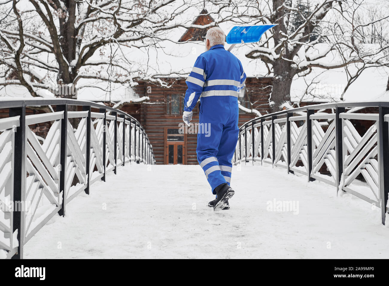 Anziani, uomo bello con la pala sulla spalla sul ponte invernale a piedi casa in legno. Concetto di pulizia manuale di neve in inverno dopo la tempesta di neve in cantiere. Foto Stock