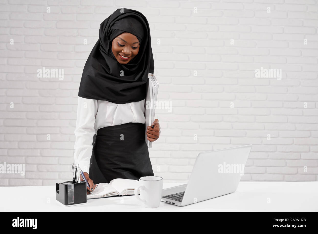Vista ritagliata della giovane donna africana vestita di nero hijab in piedi in ufficio e lavoro. Attraente femminile musulmano iscritto nel notebook e uso del computer. Concetto di multi etnico e società. Foto Stock