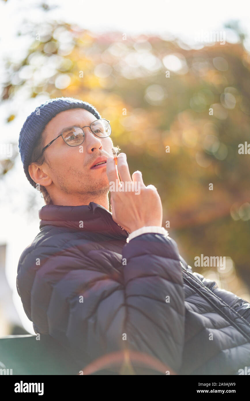 Giovane uomo in un cappuccio di fumare una sigaretta su un banco di lavoro durante una bella giornata d'autunno. Foto Stock