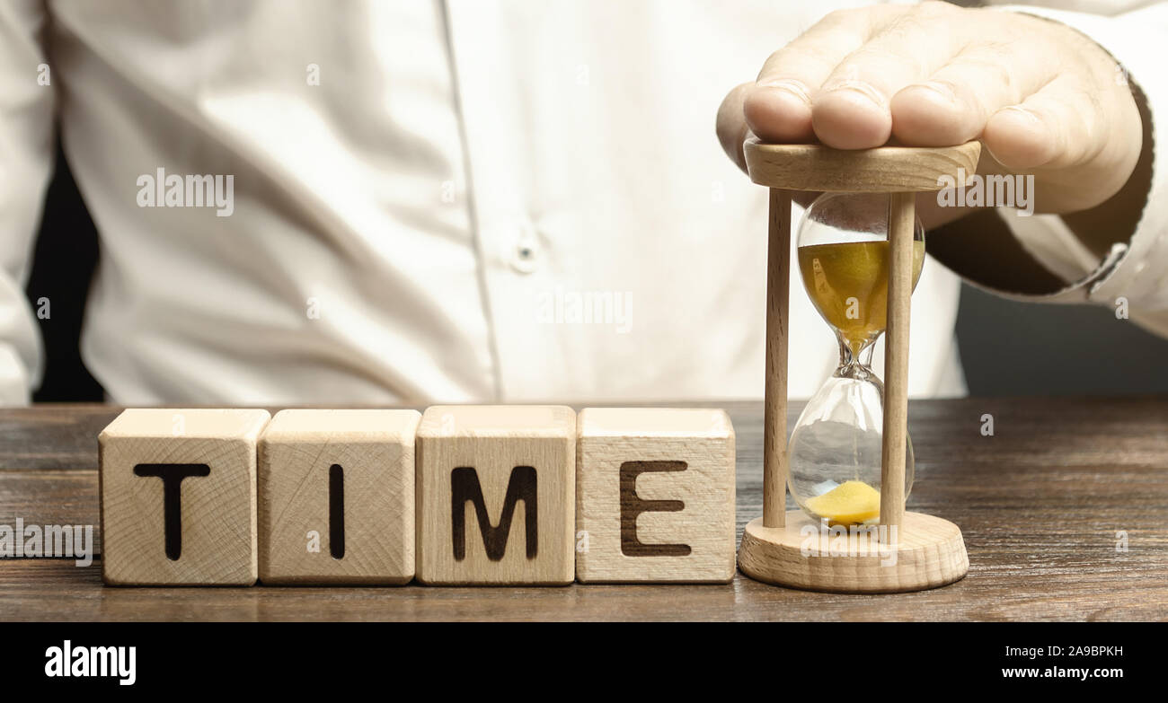 Un uomo mette la clessidra vicino a dei blocchi di legno con il tempo di parola. Concetto di gestione del tempo e la distribuzione corretta. Attività di pianificazione e di lavoro. Business y Foto Stock