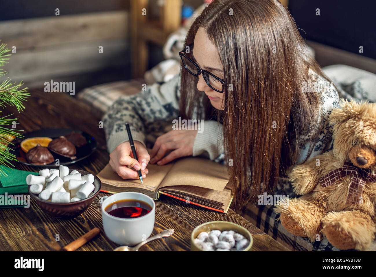 Una giovane donna in un maglione è scrivere una lista dei desideri in un notebook con una tazza di caffè la sera nella calda atmosfera del Natale. Accogliente Anno Nuovo Foto Stock