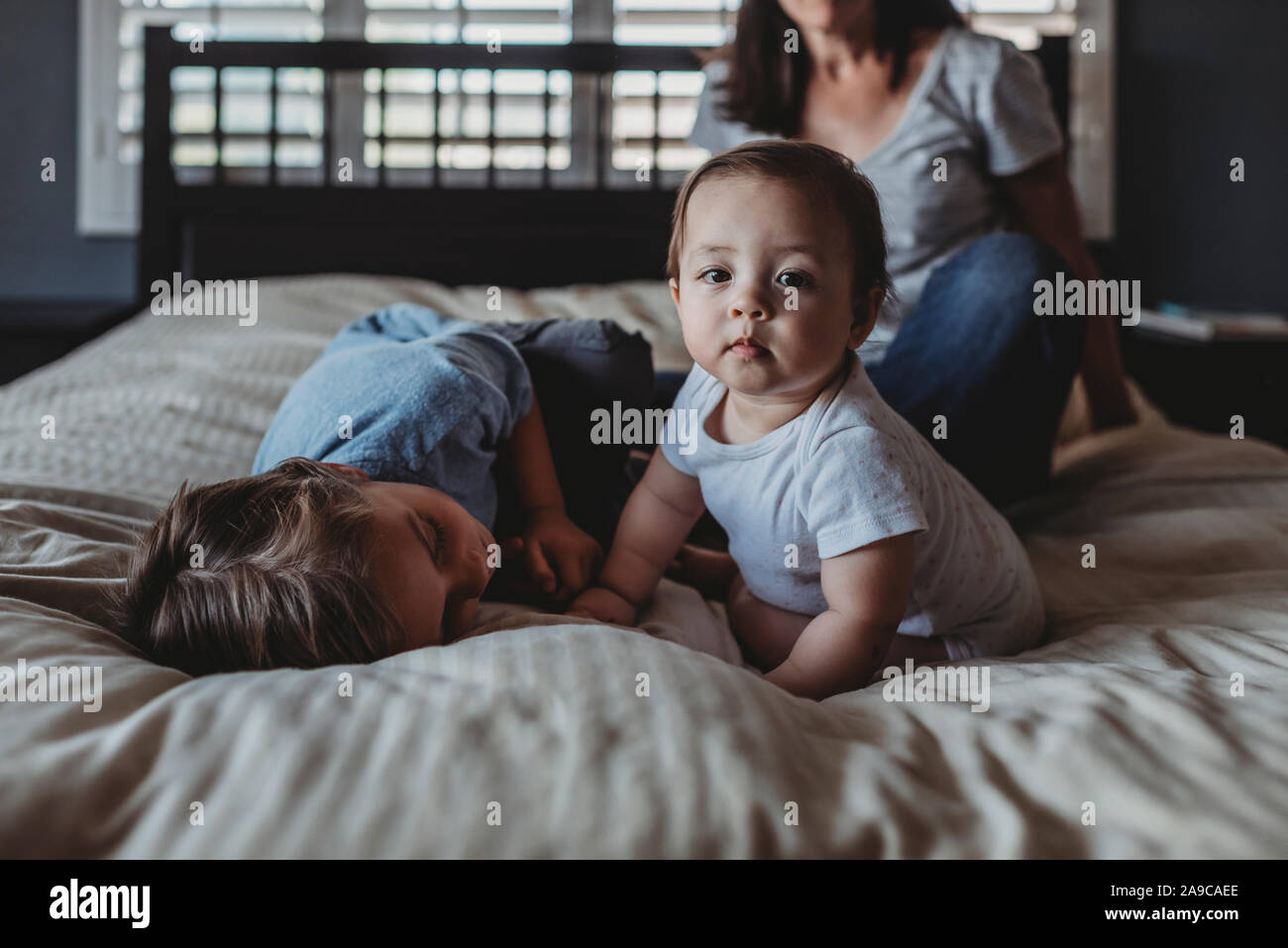 Baby ragazza seduta sul letto con mamma e 5 yr old fratello vicino a finestra Foto Stock