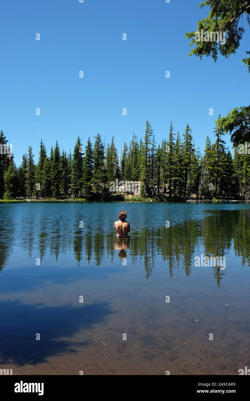 Uomo in piedi in un lago con riflessi del corpo, alberi e cielo Foto Stock